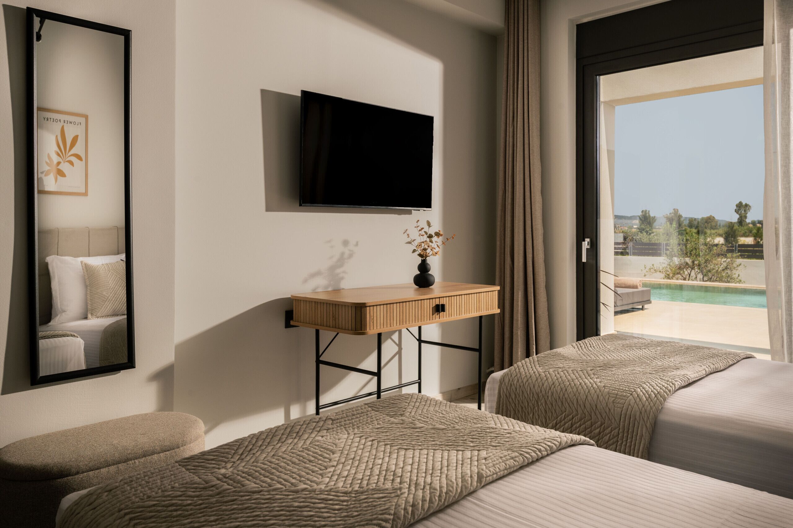 A hotel room with a bed, a bedside table with a vase, a TV mounted on the wall, and a large window with a view of a pool and greenery.