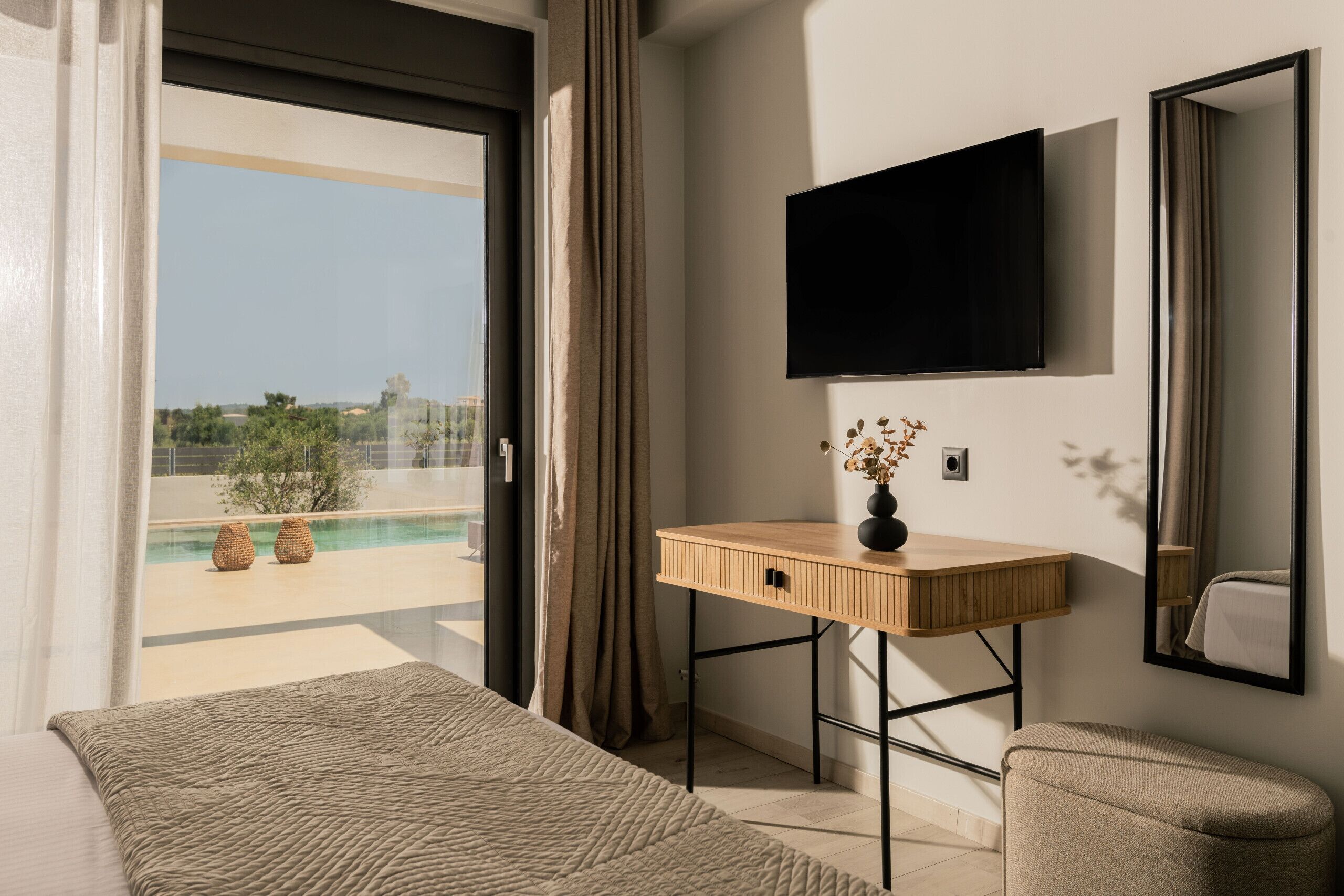 A modern hotel room with a large bed, a wooden desk, a flat-screen TV, and a view of a pool and greenery.