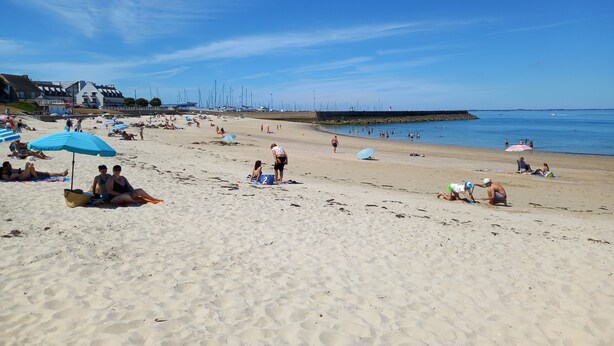 A beach with people sunbathing, umbrellas, and boats in the background.
