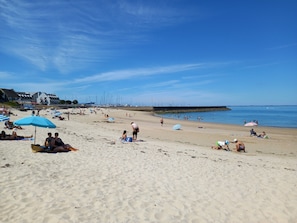 A beach with people sunbathing, umbrellas, and boats in the background.