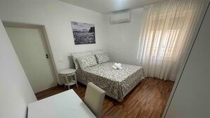 A small, clean room with a bed, a table, and a chair. There is a window with curtains and an air conditioning unit on the wall. - B&B Anna e Gaetano (Meta)