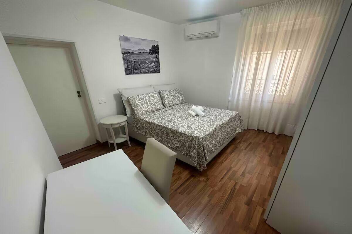 Ein kleiner, sauberer Raum mit einem Bett, einem Tisch und einem Stuhl. Ein Fenster mit Vorhängen und eine Klimaanlage sind an der Wand angebracht.