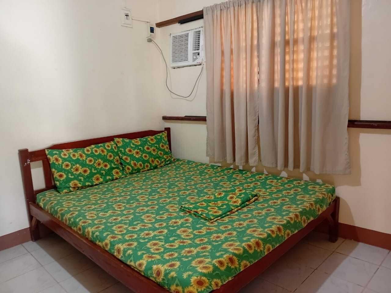 A single bed with a floral bedspread, a wooden headboard, a window with curtains, and an air conditioning unit on the wall.