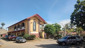 Un edificio de dos pisos con techo rojo y un gran ventanal arqueado. Varias coches están estacionados adelante, y se ve un árbol a la derecha.