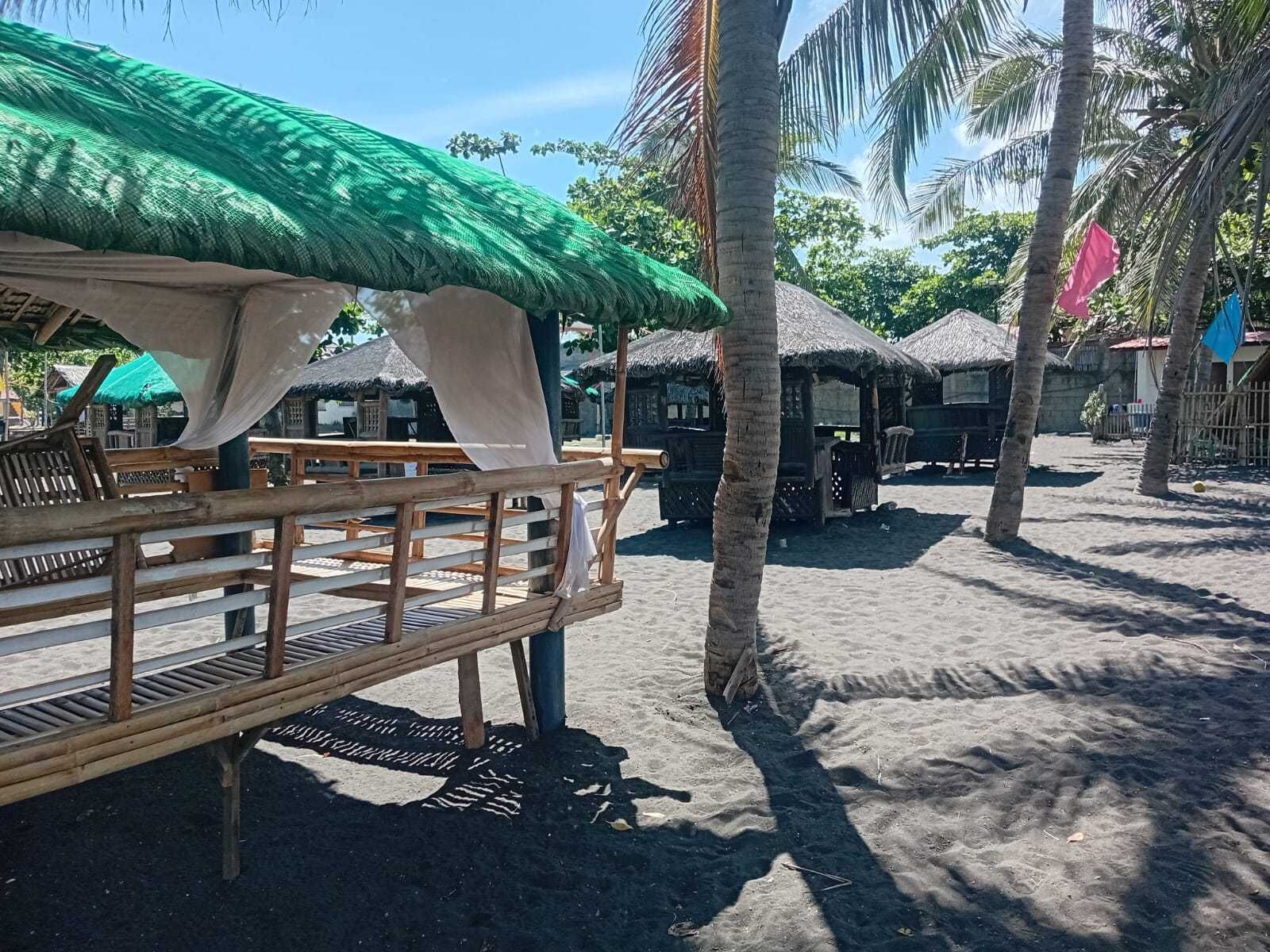 A beachside area with thatched-roof huts, wooden deck seating, and palm trees.