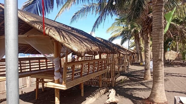 Una estructura con techo de paja y terraza de madera, rodeada de palmeras y suelo arenoso.