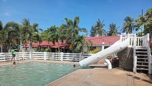 Zona de piscina con tobogán, palmeras y un edificio de techo rojo al fondo.