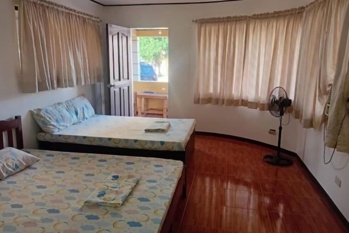 A room with two beds, a wooden floor, a fan, and a view of the outside.