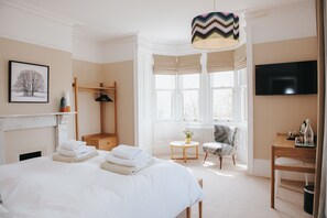 A hotel room with a bed, a wooden chair, a small round table, a radiator, a television, and a window with a view of trees.