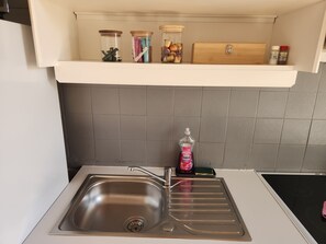 Une cuisine dotée d’un évier en acier inoxydable, d’un plan de travail blanc et d’une étagère au-dessus, où sont rangés des bocaux et une boîte en bois.