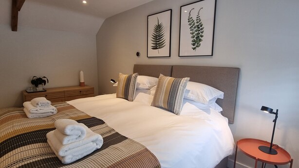 Een net opgemaakt bed met een gestreept dekbedovertrek, een nachtkastje met een lamp, en twee ingelijste botanische prints aan de muur.