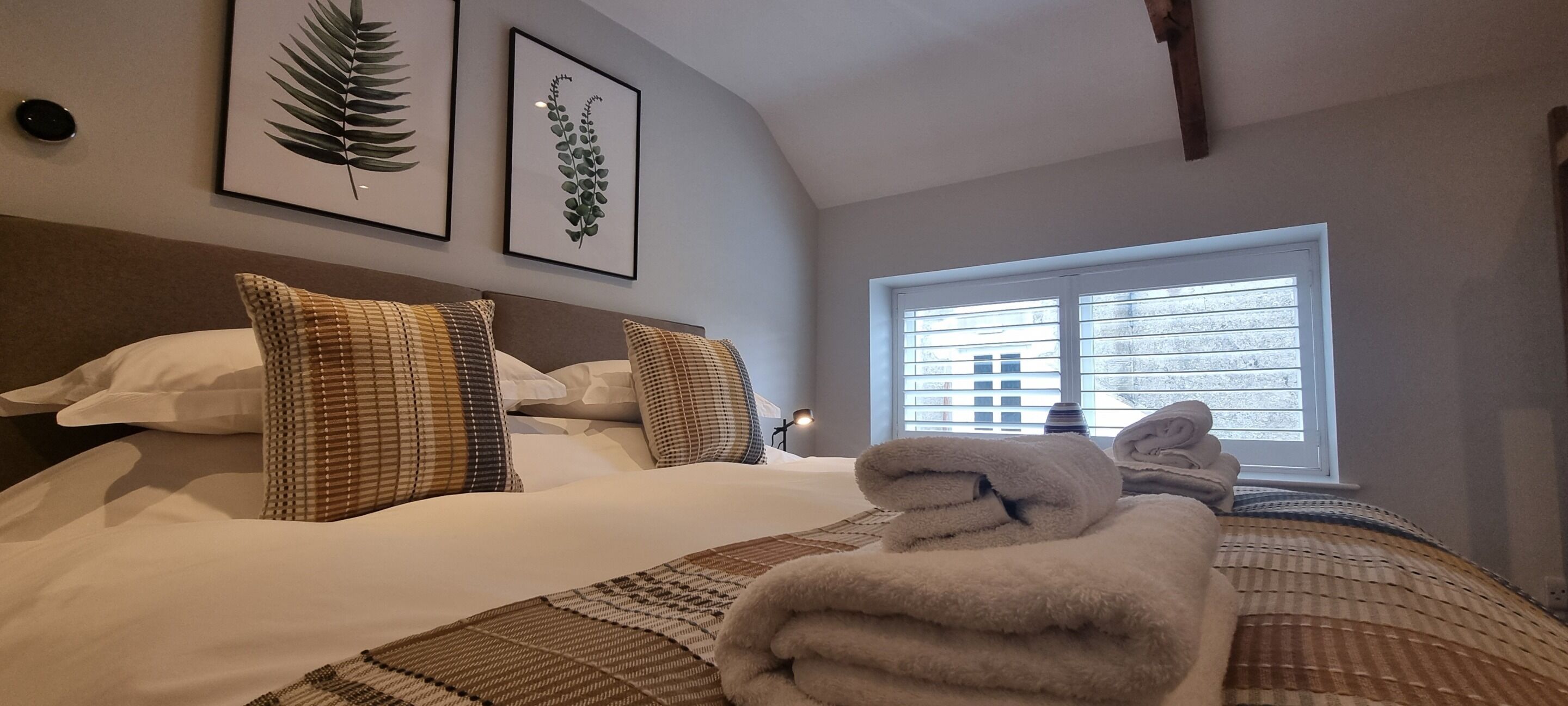 A neatly made bed with patterned pillows, a window with blinds, and framed botanical prints on the wall.