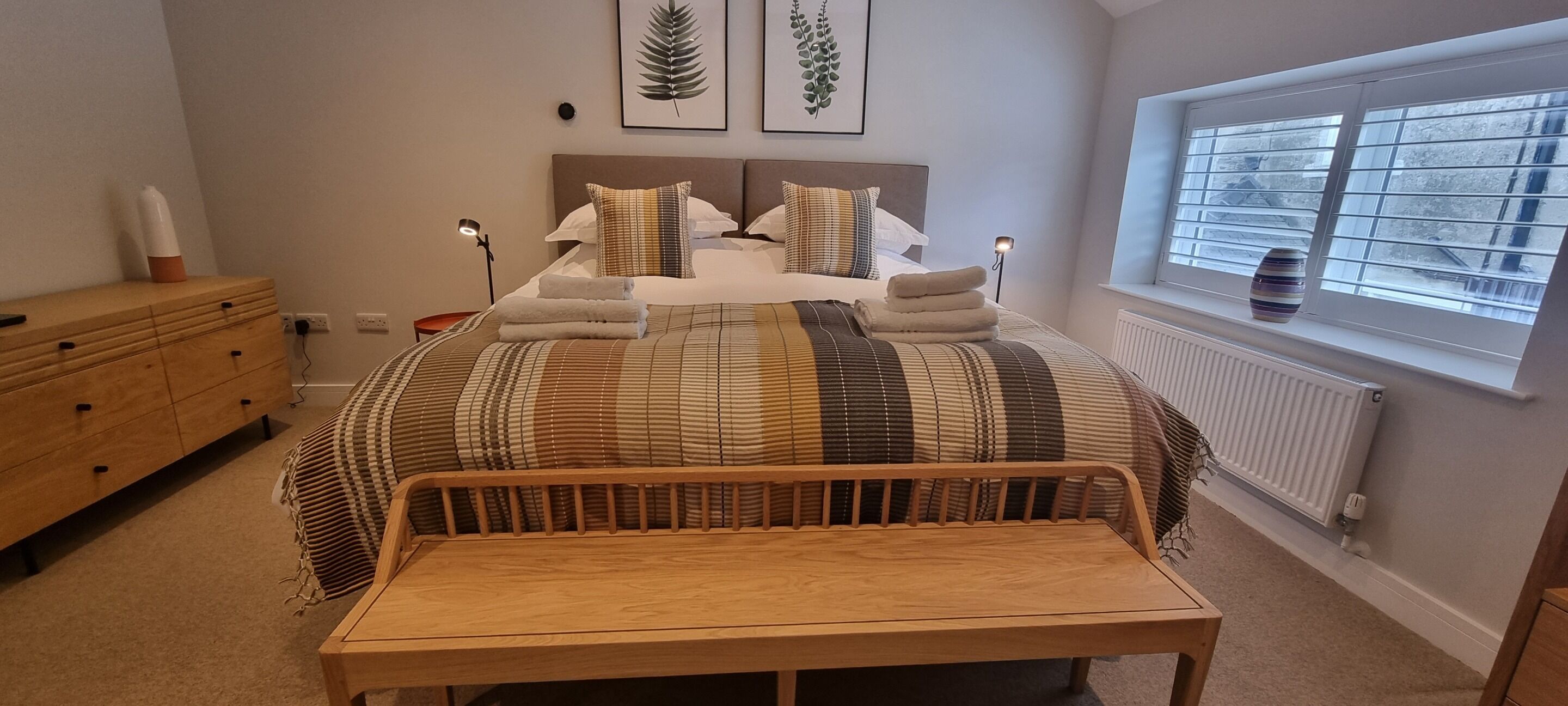Un dormitorio con cama, lámparas de noche, un banco de madera, cómoda y ventana con persianas.
