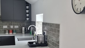 A compact kitchen with a white countertop, a red stool, a kettle, a toaster, and a clock on the wall.