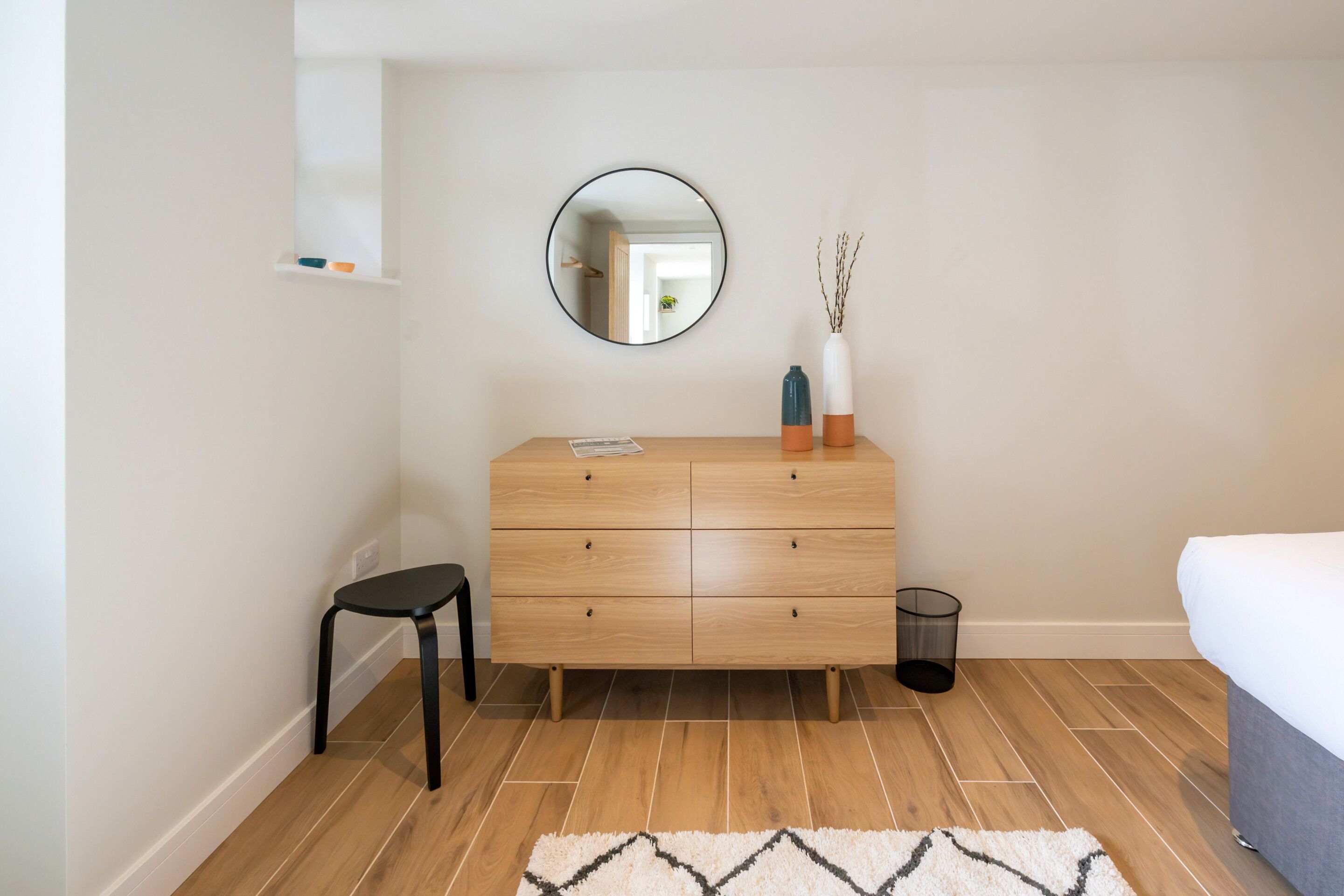 A bedroom with a wooden dresser, a round mirror, a small stool, a trash can, and a bed.