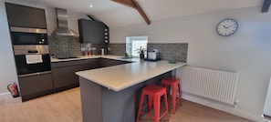 A modern kitchen with a central island, red bar stools, a built-in oven, and a clock on the wall. - The Drey Apartment - self-catered Dales luxury (Leyburn)