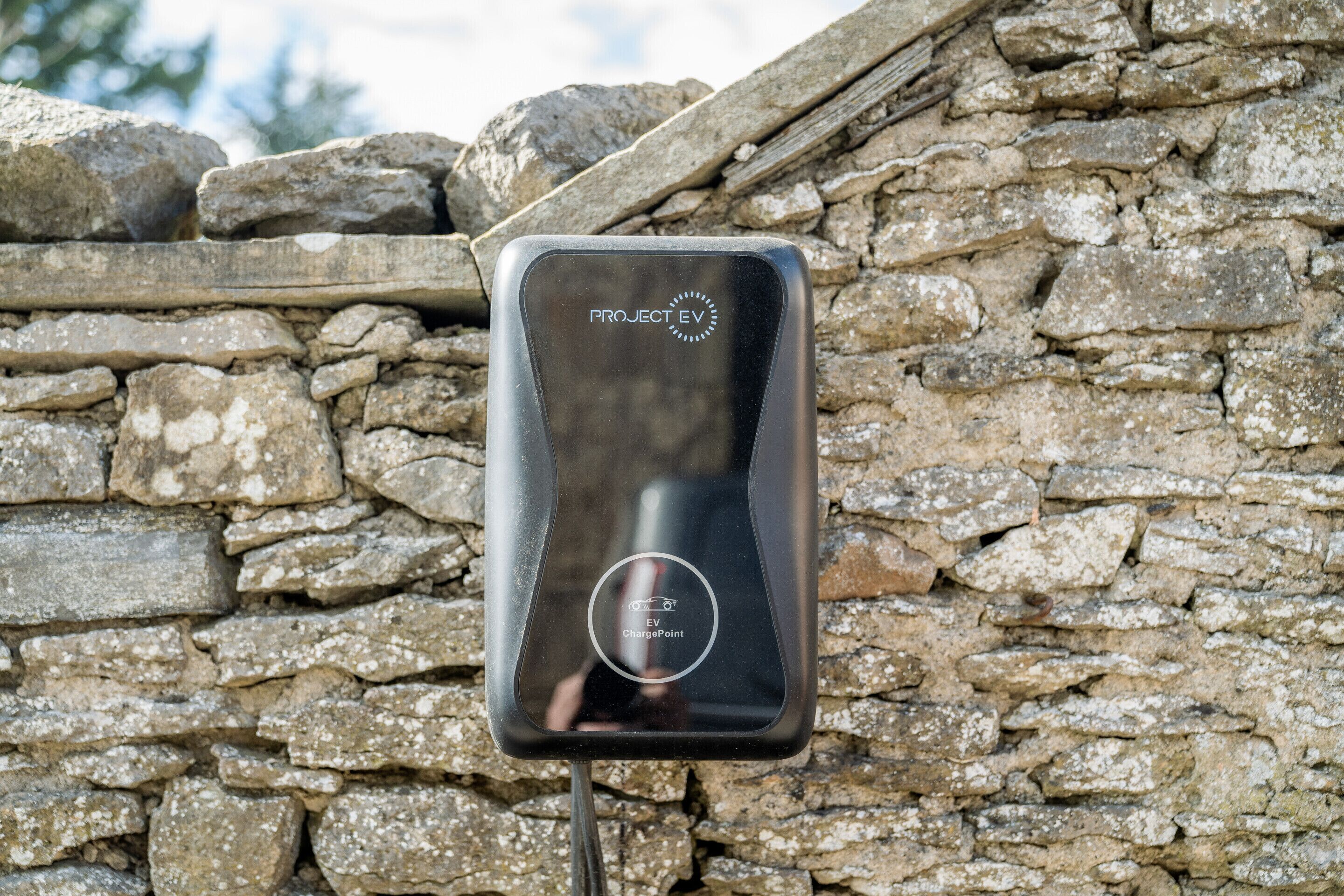 A black electric vehicle charging station labeled "PROJECT EV" against a stone wall.