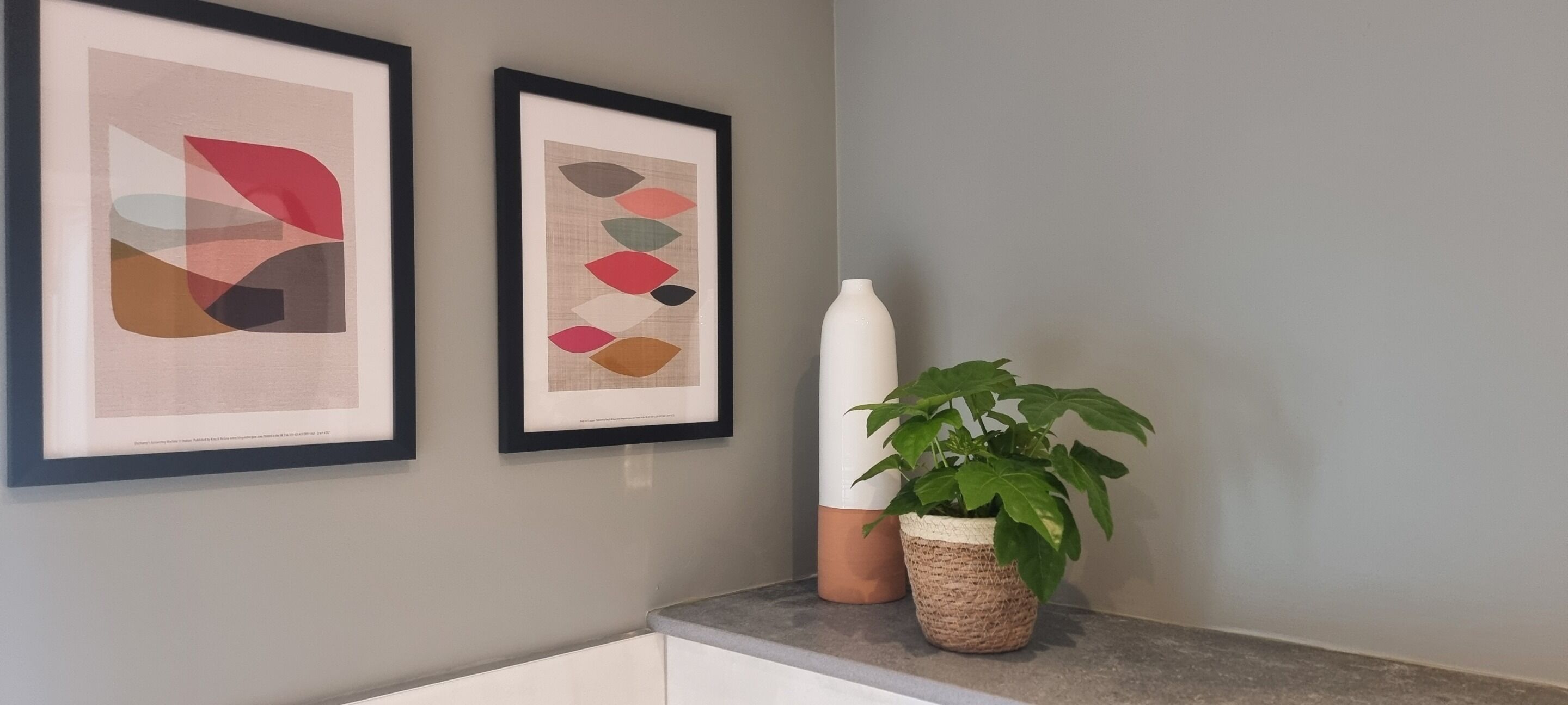 Deux œuvres d’art abstraites encadrées sont accrochées au mur, une plante en pot et un vase blanc sont posés sur un comptoir.