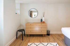Une chambre avec une commode en bois, un miroir rond, un petit tabouret, une poubelle et un lit.