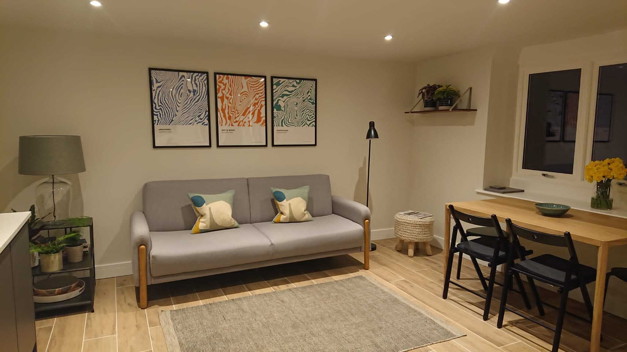 Un salón moderno con un sofá gris, dos sillones, una mesita y una lámpara de pie. Se observan tres obras de arte enmarcadas en la pared.