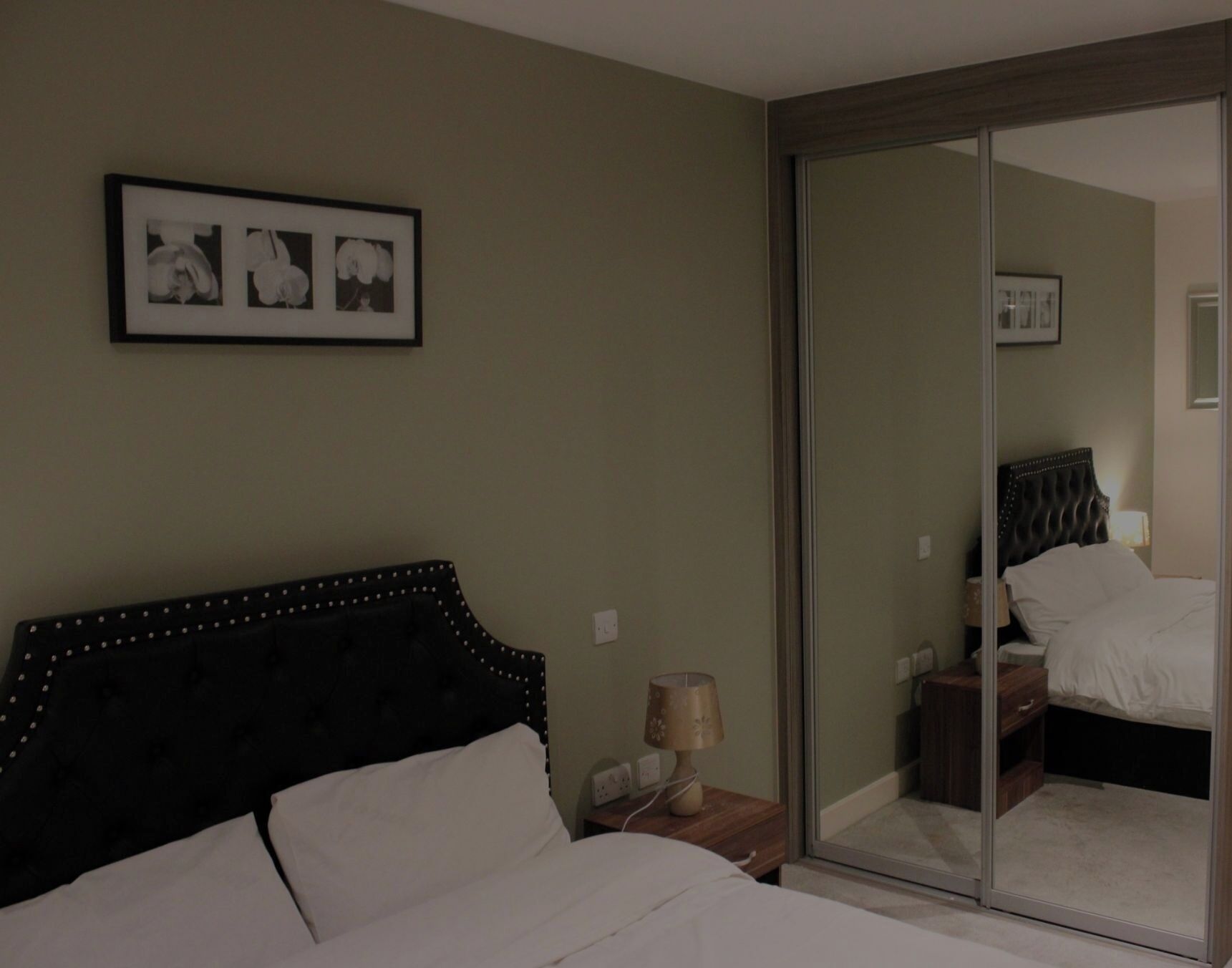 Een slaapkamer met een bed, nachtkastje, lamp en een lijst aan de muur.