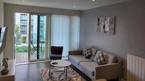 Ein modernes Wohnzimmer mit einer Couch, einem kleinen runden Tisch und einem Sessel. Es gibt ein großes Fenster mit Jalousien, ein Bild an der Wand und eine Heizung.