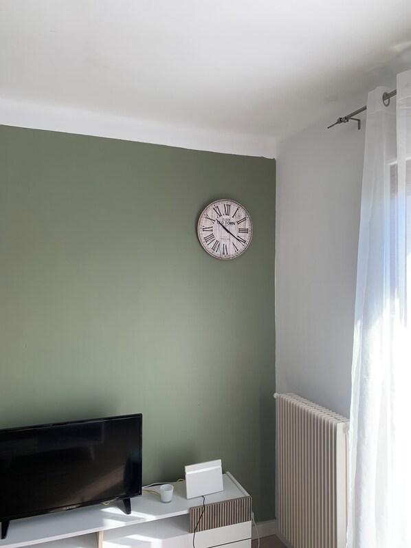 Une pièce avec un mur vert, un radiateur blanc, un téléviseur à écran plat noir et un thermostat blanc fixé sur une armoire blanche.