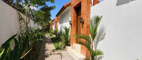 A pathway leading to a building with a brick facade and white walls, surrounded by lush greenery and a clear blue sky.