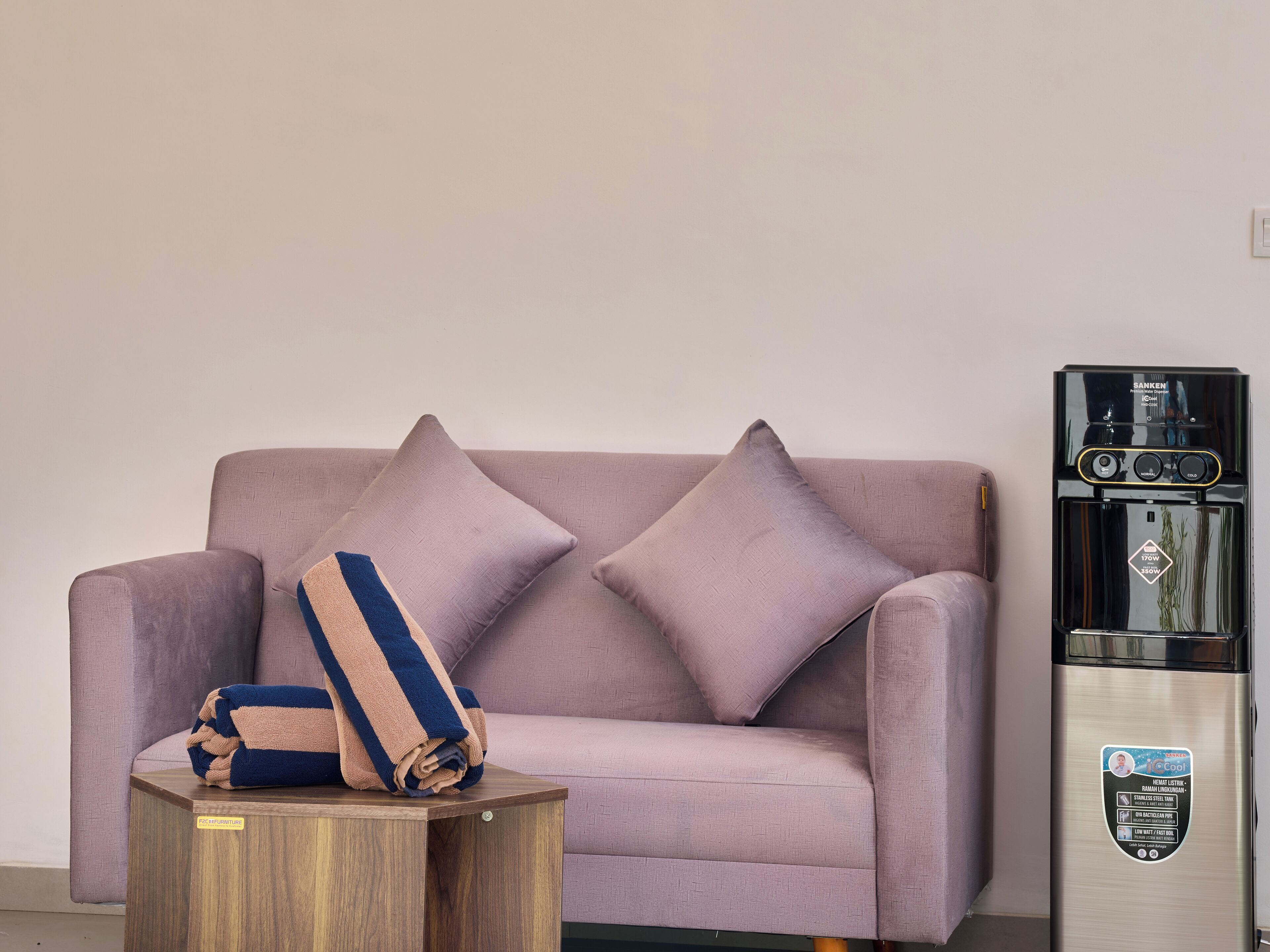 A living room with a sofa, two pillows, a small wooden side table, and a stainless steel water dispenser.