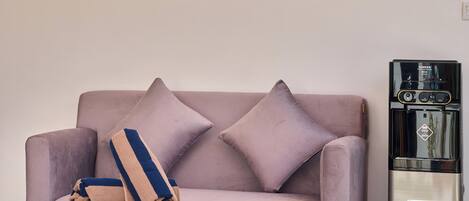 A living room with a sofa, two pillows, a small wooden side table, and a stainless steel water dispenser.