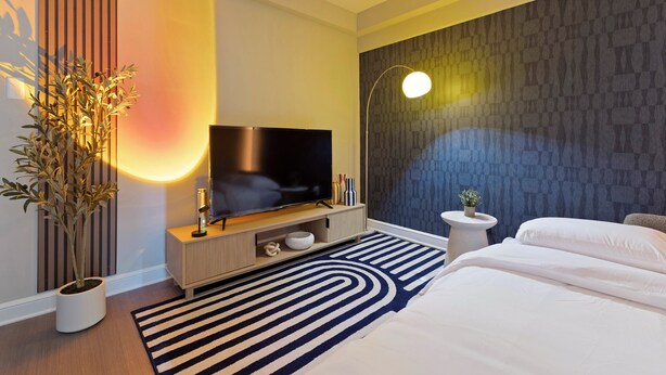 A modern hotel room with a flat-screen TV, a potted plant, a striped rug, and a bed with white linens. - Arlington 1BR • Metro Access • Free Parking (Arlington)