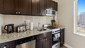 A modern kitchen with dark brown cabinets, a granite countertop, stainless steel appliances including a microwave, toaster, and dishwasher, and a window with a city view. - Arlington 1BR • Metro Access • Free Parking (Arlington)
