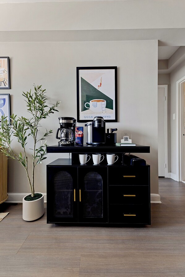 A modern kitchen area with a coffee station, a potted plant, and a framed artwork on the wall.