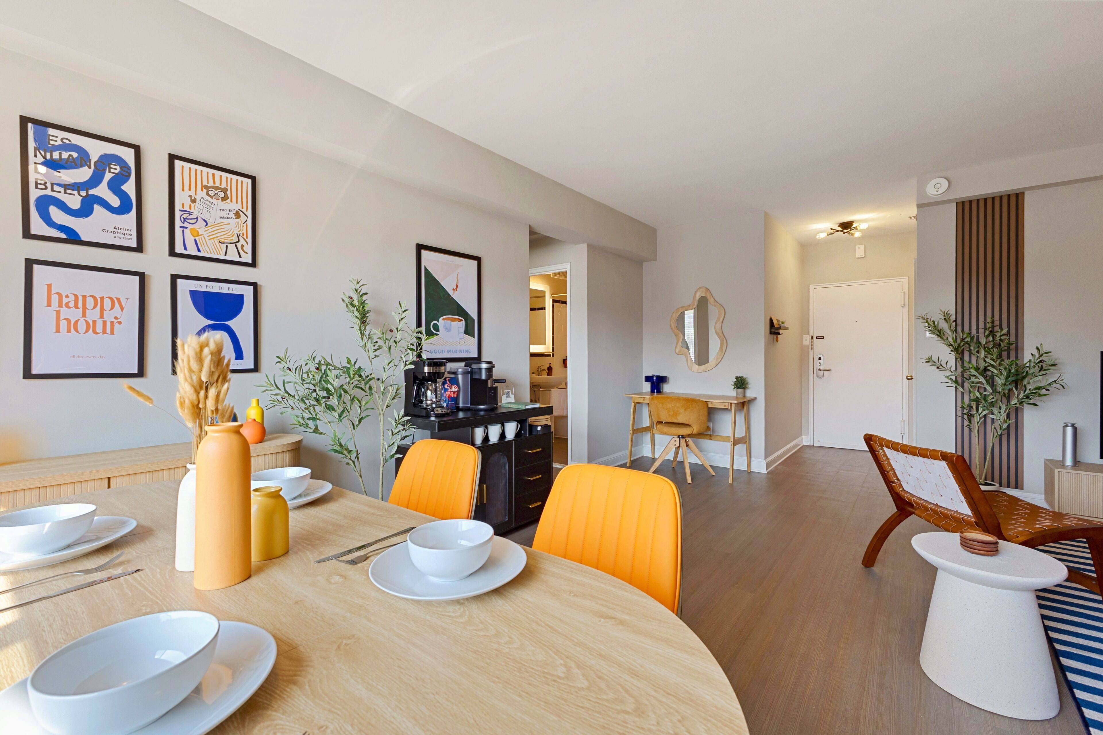 Un comedor moderno con una mesa de madera dispuesta para cuatro personas, rodeada de sillas color naranja. Hay cuadros enmarcados en la pared, una cafetera y una pequeña planta en la mesa.