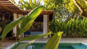 Un área de piscina con terraza de madera, estructura de techo de paja y exuberante vegetación tropical.
