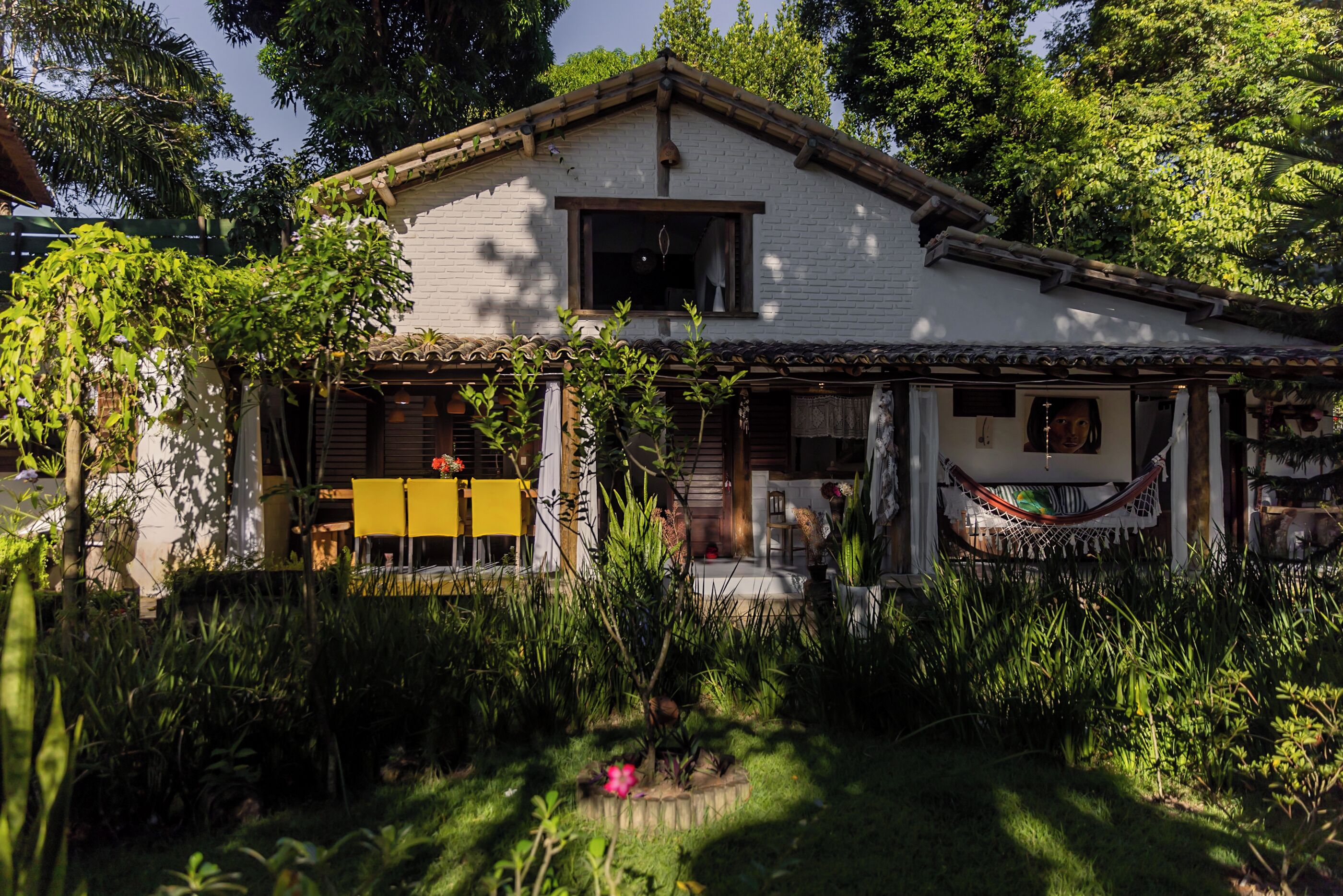 A white house with a tiled roof, surrounded by lush greenery and a hammock on the porch.