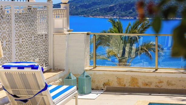 A balcony with a white chair, a small table, and a view of the sea and hills.