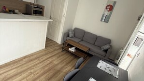 Ett kompakt bostadsutrymme med ett litet kök, en matplats med bord och stolar, en grå soffa och ett litet soffbord.