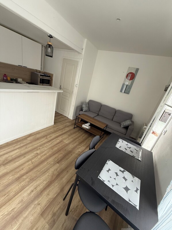 A compact living space with a kitchenette, a dining area with a table and chairs, a gray sofa, and a small coffee table. - Charming townhouse, fully equipped, 2 steps from shops (Grand-Couronne)