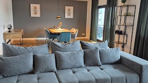 Een moderne woonkamer met een grijze hoekbank, een eettafel met stoelen, een klein rond tafeltje en een boekenkast.