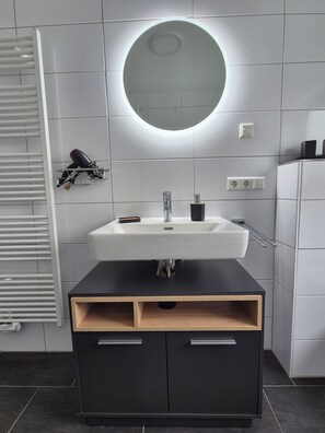 Un baño moderno con un lavamanos blanco, tocador negro y un espejo redondo con luz superior.