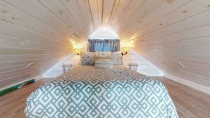Un dormitorio con techo inclinado, una cama con ropa de cama estampada, dos lámparas de noche y una ventana con cortinas.