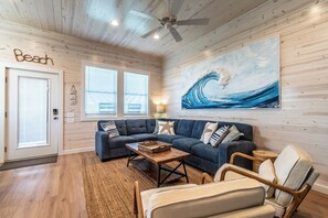 A living room with a dark blue sofa, wooden flooring, a ceiling fan, and a large wave painting on the wall. - Luxury 4-Bedroom Beach Retreat | Private Pool, Boardwalk Access for Golf Carts (Port Aransas)