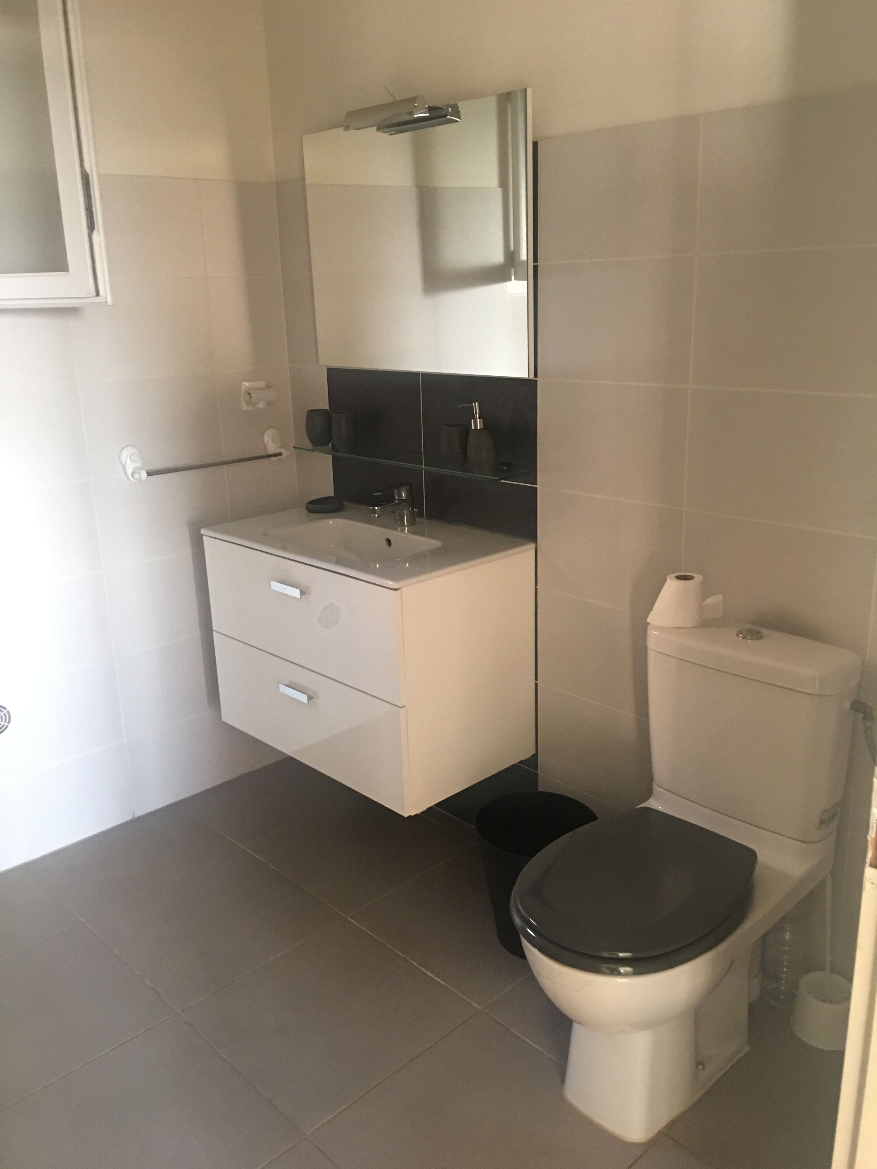 Ein modernes Badezimmer mit einem weißen Waschtisch, einer Glasablage, einer Toilette und einem Spiegel über dem Waschbecken.