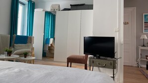 A hotel room with a bed, a TV on a metal stand, a chair, a small table, and a wardrobe.