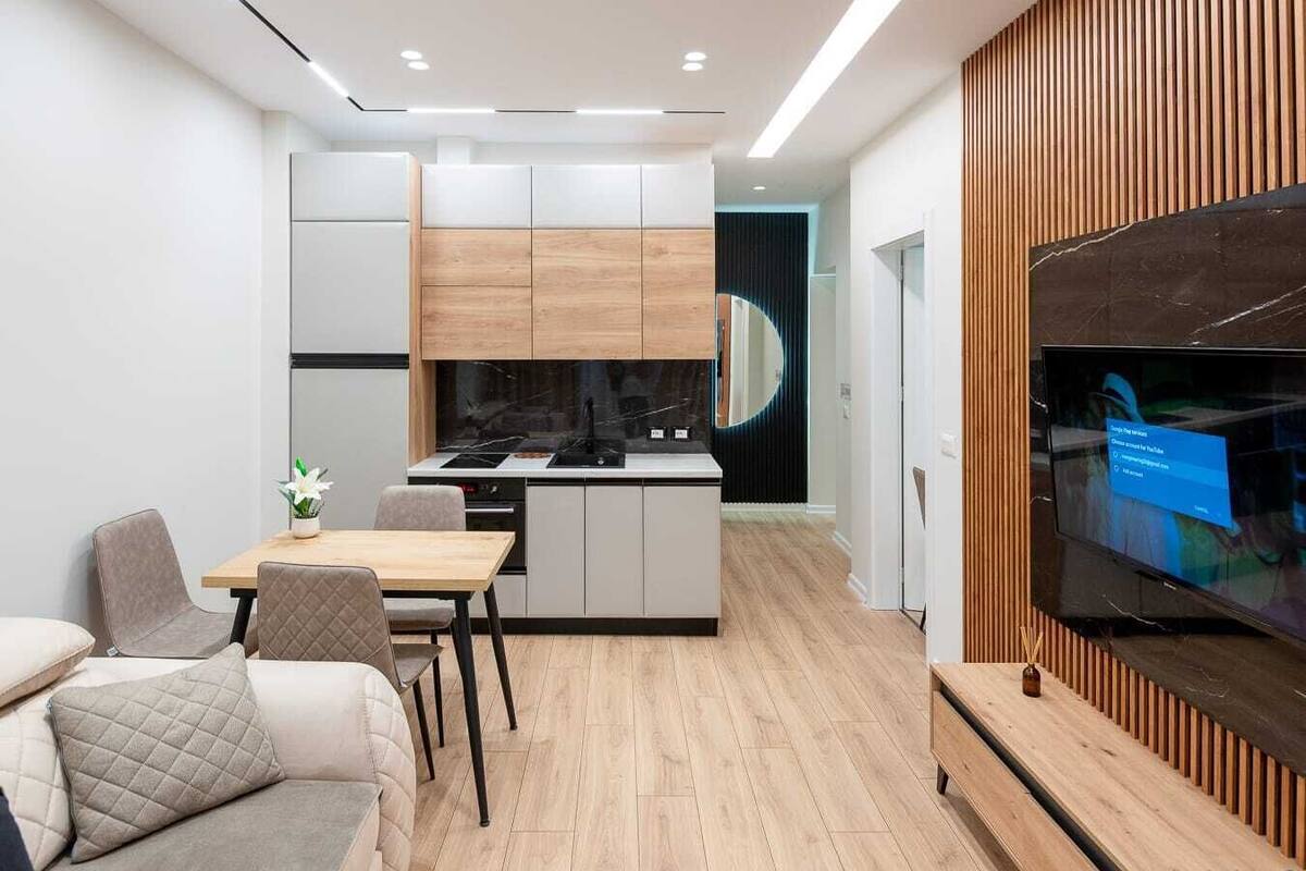 Eine moderne Küche mit einer zentralen Kochinsel, einem Essbereich mit einer Couch und Stühlen sowie einem Fernseher, der an einer Wand aus Holzpaneelen befestigt ist.