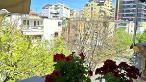 A balcony with a view of city buildings, a wooden table, and red flowers.