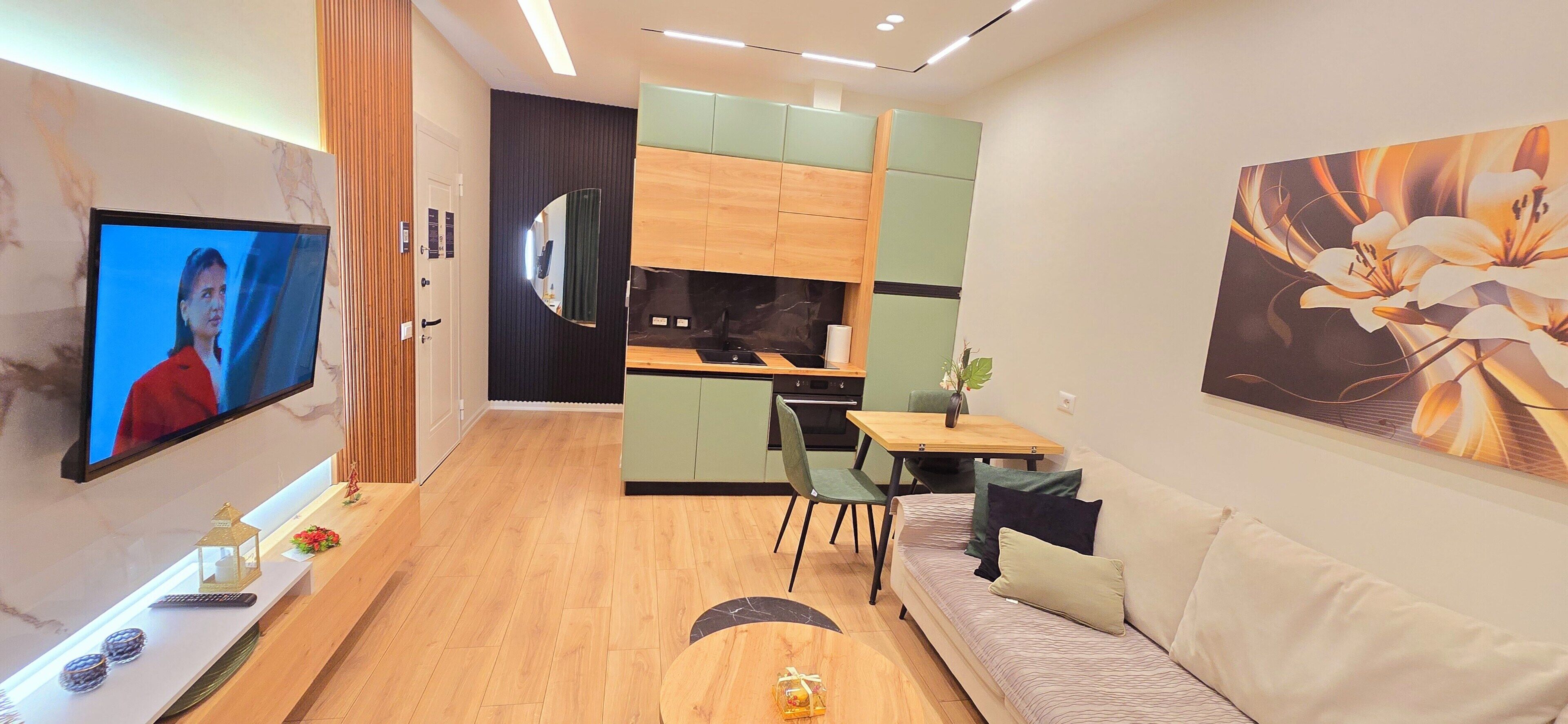 A modern living room with a flat-screen TV, a sofa, a dining table, and a kitchen area with green cabinets.