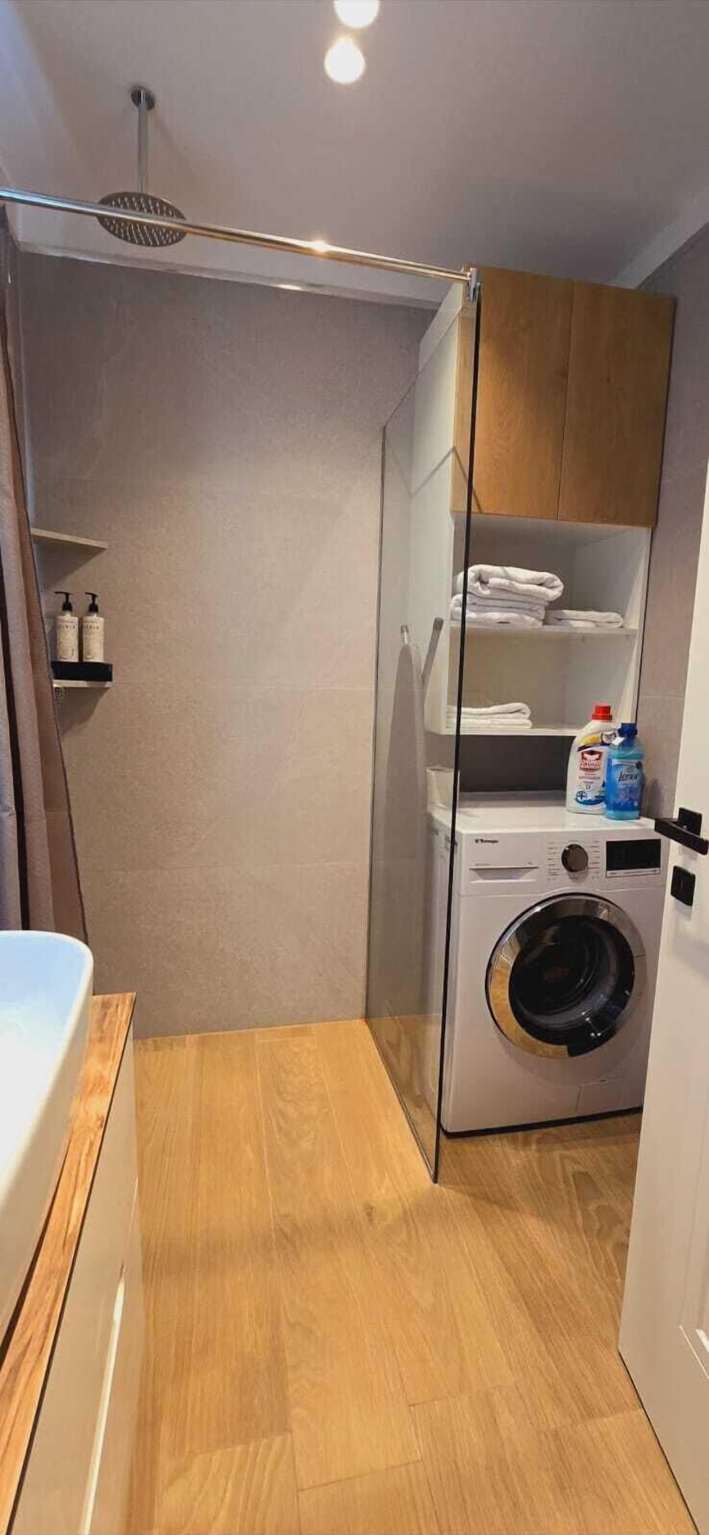 A compact bathroom with a shower, a washing machine, and a shelf with toiletries.