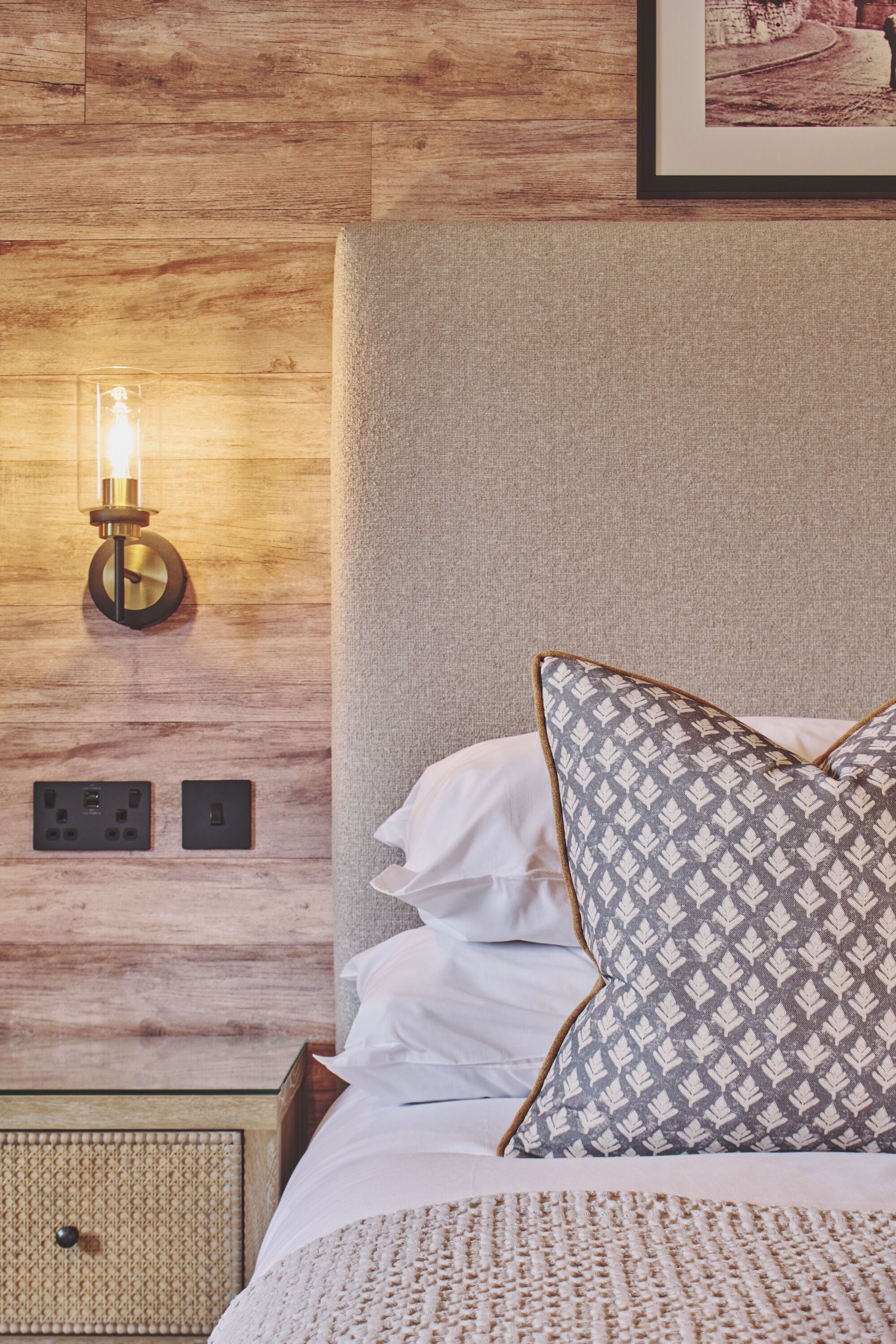 A neatly made bed with patterned pillows, a wall-mounted light fixture, and a framed picture above the bed.